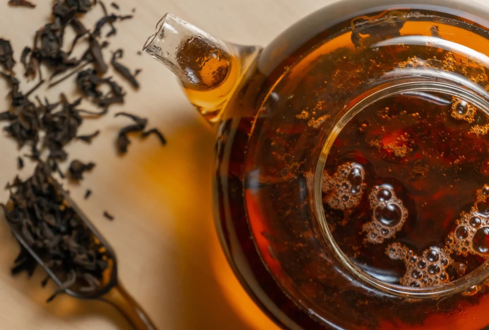 Dark tea leaves beside a scoop, with a glass teapot of brewed tea seen from above