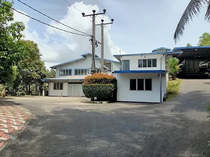 A white two-storey factory building with a blue roof, seen across a paved yard with trees around it