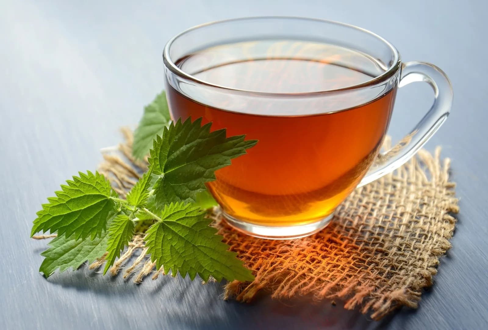A clear glass cup of amber tea on a square of woven burlap, with fresh green leaves beside it